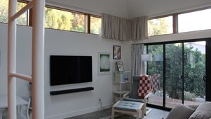 Een moderne woonkamer met een flatscreen televisie aan de muur, een geruit fauteuil, een bank en een eethoek met een tafel en stoelen.