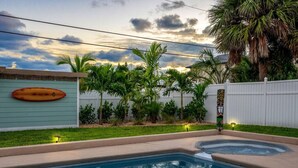 A swimming pool with a curved edge, surrounded by a white fence, palm trees, and a surfboard decoration on a wall.