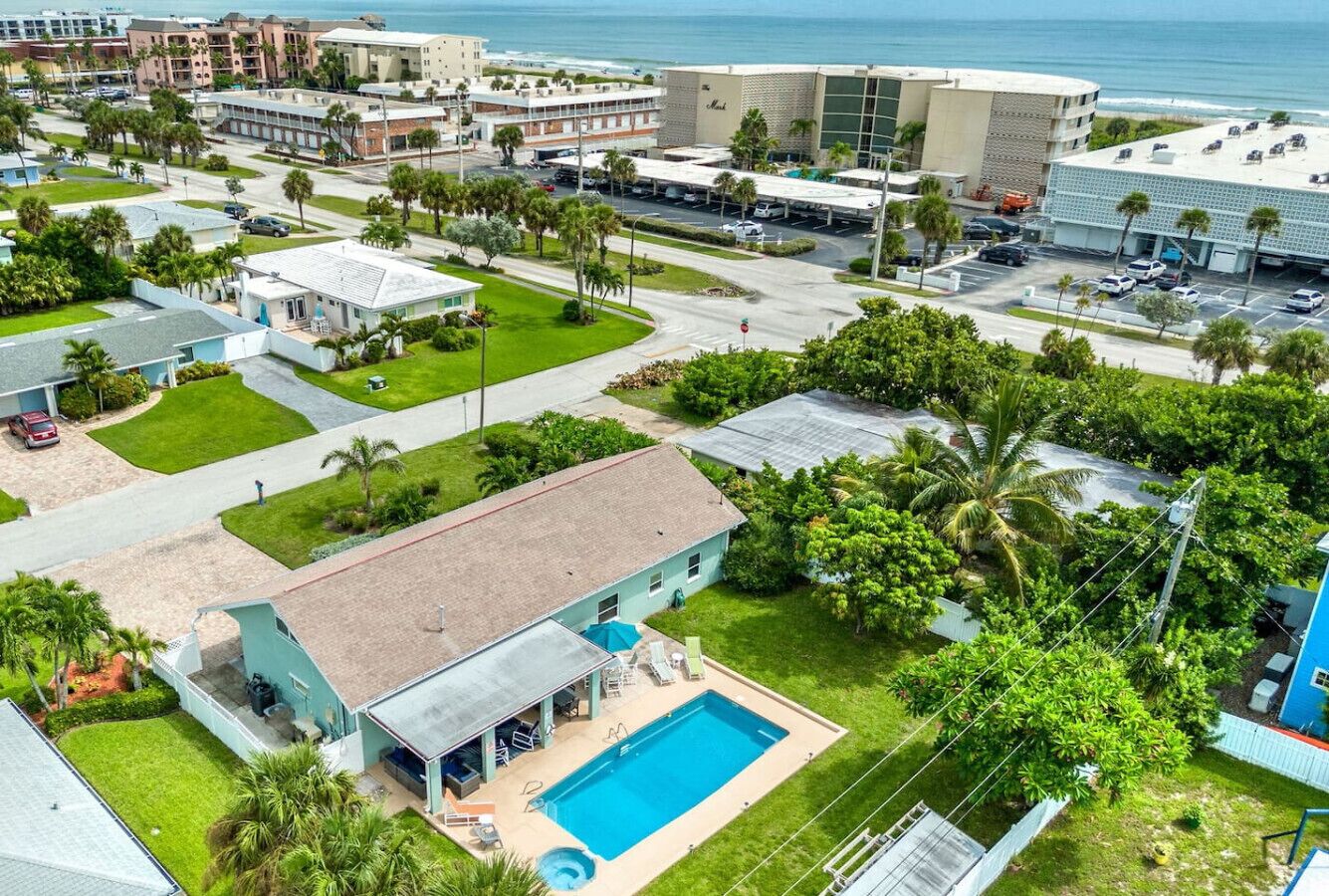 An aerial view of a residential area with a house featuring a swimming pool, surrounded by greenery and a view of the ocean.