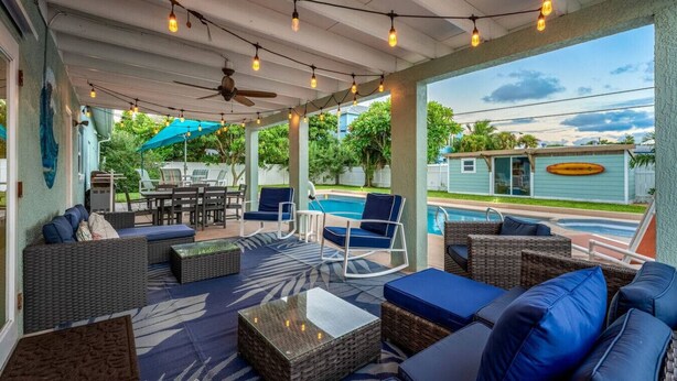 A covered outdoor area with wicker furniture, a ceiling fan, string lights, and a view of a swimming pool and house.