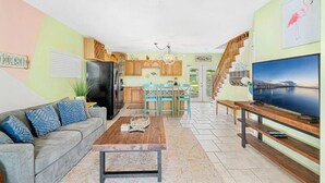 A living room with a sofa, coffee table, and TV. There is a kitchen with wooden cabinets and a dining area with chairs.