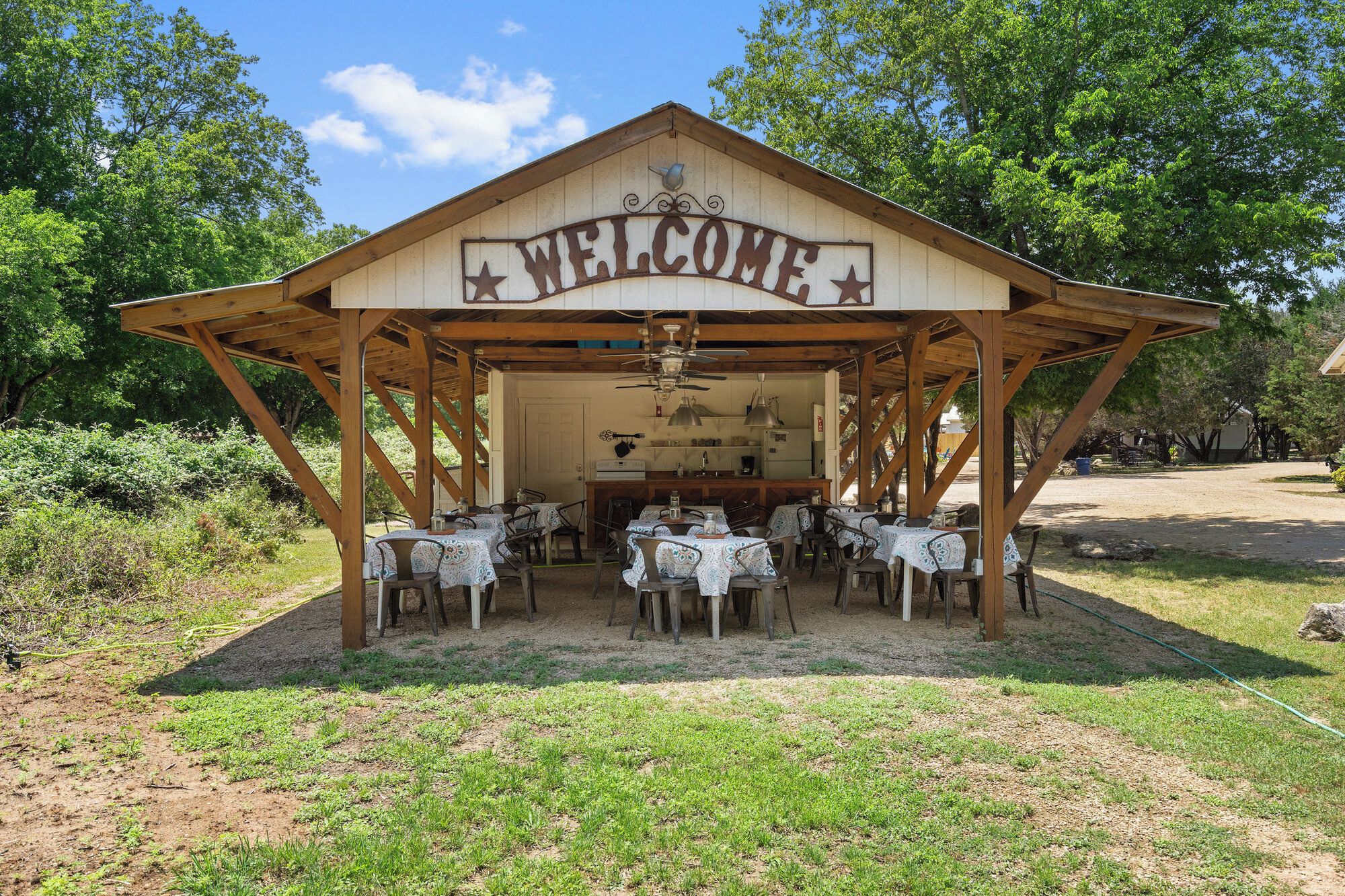 A wooden pavilion with a 'WELCOME' sign, tables with white tablecloths, and a kitchen area.