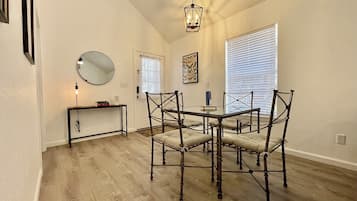 A dining area with a round table and chairs, a hanging light fixture, a mirror, and a window with blinds.