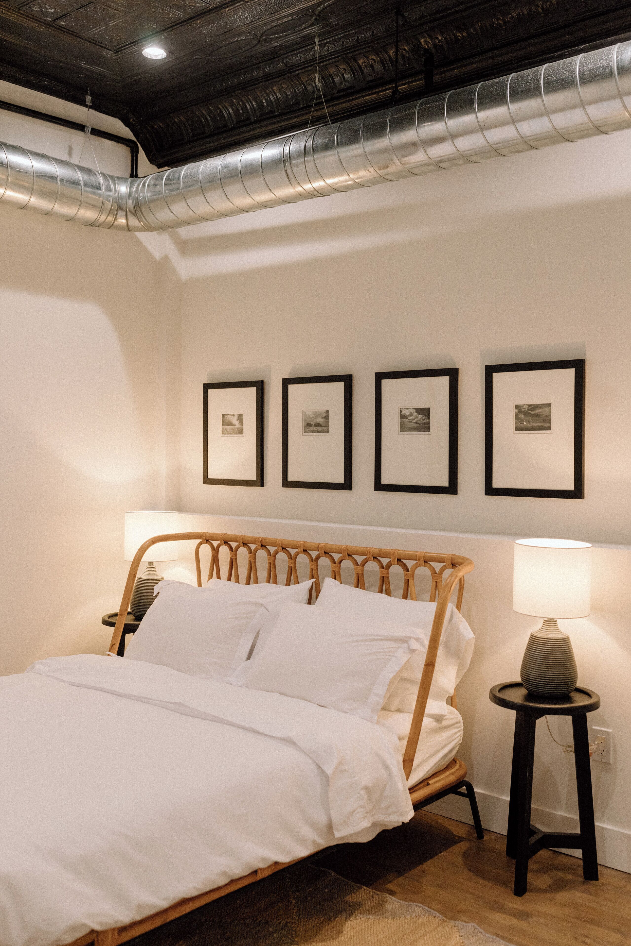 A neatly made bed with white linens, a wooden headboard, and a bedside table with a lamp. There are four framed pictures on the wall above the bed.