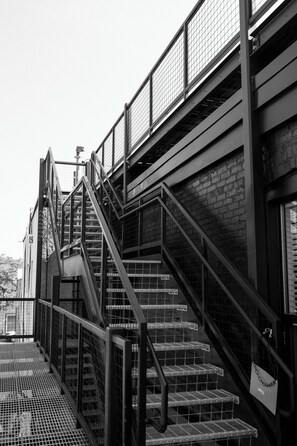 Vista en blanco y negro de una escalera de metal con barandilla, que conduce a un muro de ladrillo.