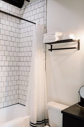 Un baño con paredes de azulejos blancos, una bañera negra, un inodoro blanco y un perchero con toallas dobladas.