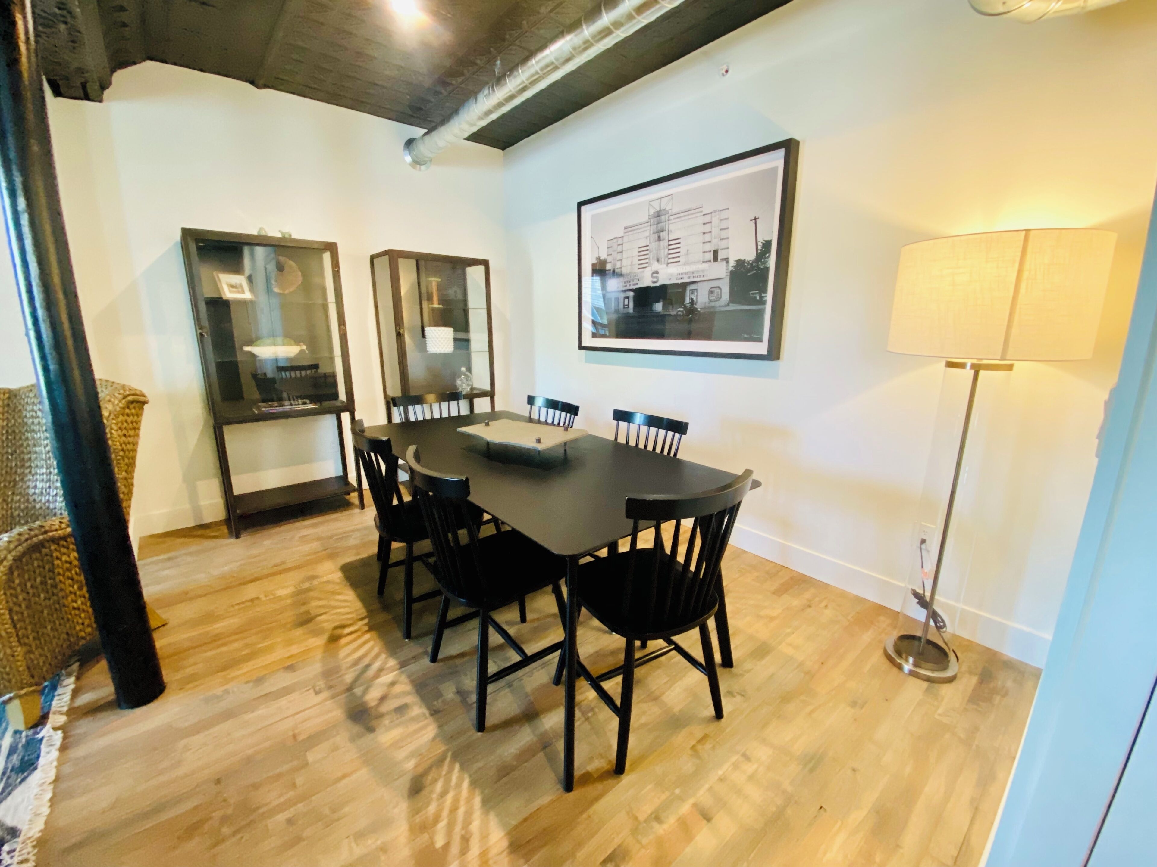 Un coin repas avec une table rectangulaire entourée de chaises, un tableau encadré au mur et un lampadaire.