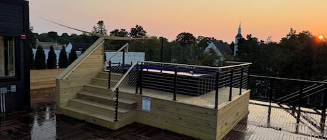 Um deck de madeira com piscina, escadas e vista para uma torre de igreja ao fundo.