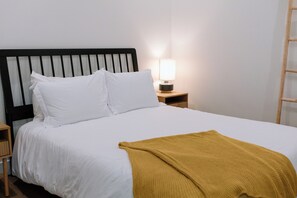 Una cama con ropa de cama blanca, un plaid color mostaza, una mesita de noche de madera con una lámpara y una escalera de madera apoyada en la pared.