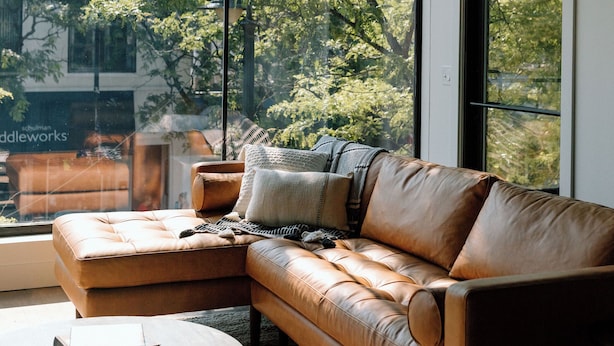 A modern living room with a leather sofa, a round coffee table, and a large window offering a view of trees and a building. - The Kroger Lofts (Charlevoix)