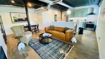 A modern living room with a brown leather sofa, a round coffee table, and a patterned rug. There is a dining area with a black chair and a table, and a kitchen in the background.