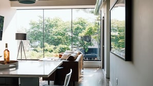 A modern interior with a dining area, a sofa, and large windows offering a view of trees and a building. - The Kroger Lofts (Charlevoix)