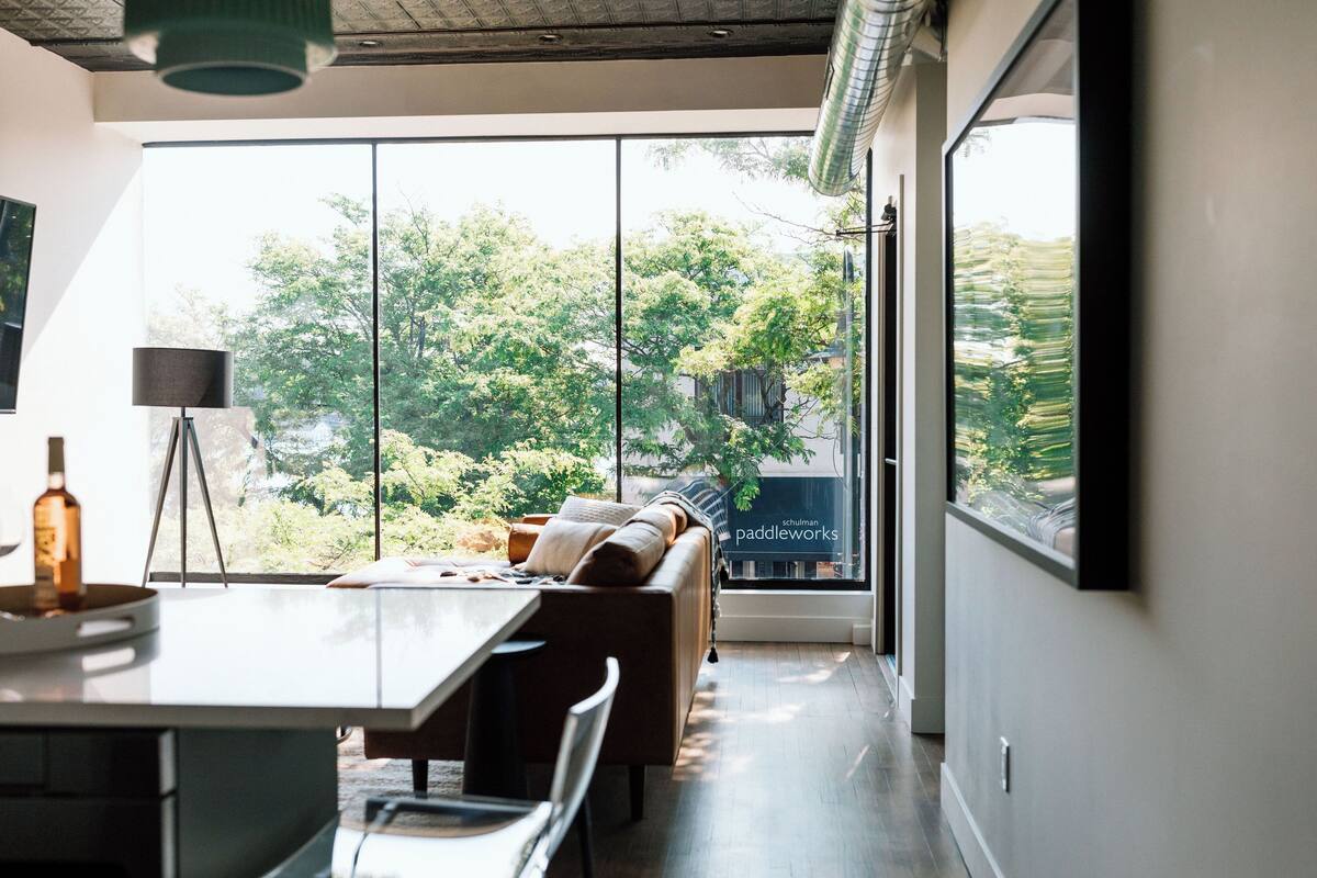 Un interior moderno con comedor, sofá y amplios ventanales con vistas a árboles y un edificio.