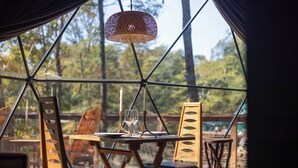 Comedor con mesa y sillas, una lámpara colgante y vista a árboles a través de una estructura de ventana geométrica.