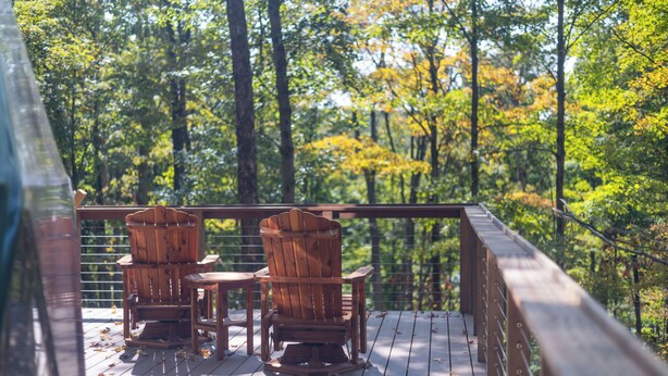 Una terraza de madera con dos sillas Adirondack y una mesa pequeña, rodeada de un bosque.