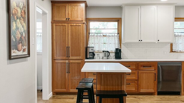A modern kitchen with wooden cabinets, a white countertop, and a stainless steel dishwasher. There are two stools and a window with a view of greenery. - The Gem - Spacious 3/2 House near ACU (Abilene)