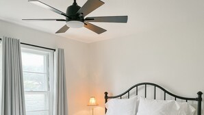 A neatly made bed with white linens, a ceiling fan, a bedside lamp, and a window with curtains. - The Gem - Spacious 3/2 House near ACU (Abilene)