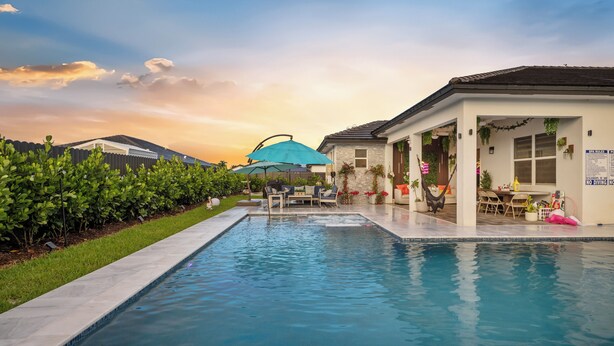 Piscina con zona de patio, espacio de estar techado y vista a un barrio residencial al atardecer.