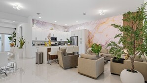 Una cocina moderna con armarios blancos, electrodomésticos de acero inoxidable y salpicadero de mármol. Hay una zona de comedor con sillas blancas y una mesa, y una sala de estar con sofás y una mesa de centro.