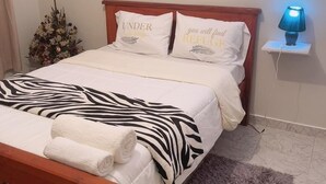 Een net opgemaakt bed met een deken met zebraprint, twee kussens met tekst, een kleine bedlamp en een houten nachtkastje.
