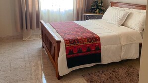 Een slaapkamer met een bed, een houten hoofdbord, een nachtkastje, een kleine tafel met een plant en een raam met gordijnen.