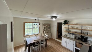 Une cuisine avec une table à manger en bois, des chaises blanches, un four encastré et des étagères remplies d’assiettes et de verres.
