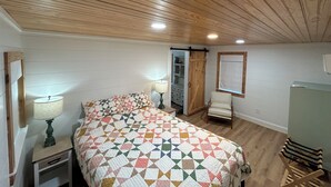 Une chambre avec un plafond en bois, un lit recouvert d’une couverture aux couleurs vives, une table de chevet avec une lampe, une chaise et une armoire.
