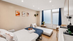 Ein Hotelzimmer mit zwei Betten, einem kleinen Tisch mit Blumenvase, einem Fenster mit Vorhängen und einer Küchenzeile.