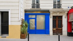 Ein Gebäude mit einer blauen Tür, auf der 'COIFFURE' steht, einer Holztür mit schmiedeeisernem Gitter und einem Blumentopf auf dem Bürgersteig.