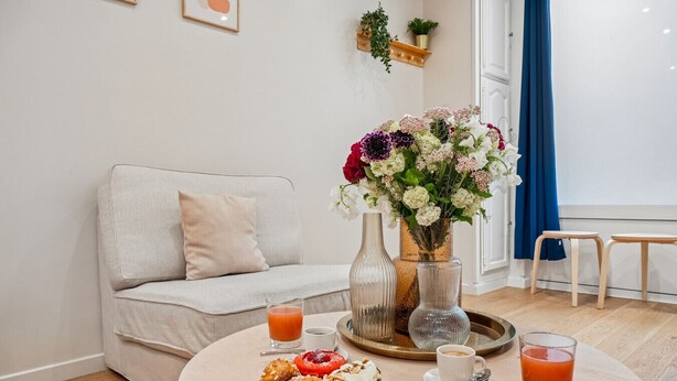 Ein Zimmer mit einer Couch, einem runden Tisch mit Speisen und Getränken, einer Vase mit Blumen und einem Fenster mit Vorhängen.