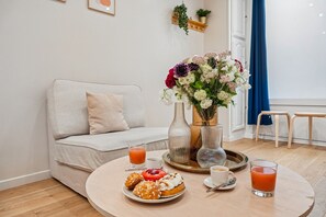 Une pièce avec un canapé, une table ronde sur laquelle sont disposés de la nourriture et des boissons, un vase rempli de fleurs et une fenêtre avec des rideaux.