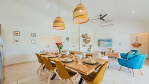 A dining area with a wooden table set for six, surrounded by yellow chairs. There are hanging wicker lamps above the table, a ceiling fan, and a wall-mounted TV.