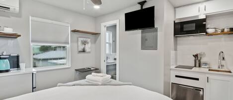 A modern hotel room with a bed, a ceiling fan, a TV, a kitchenette, and a window with blinds.