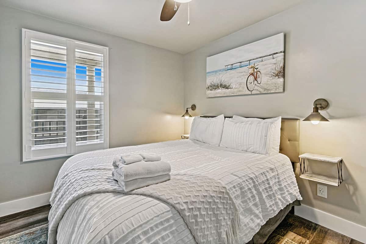 A bedroom with a large bed, two bedside lamps, a wall-mounted shelf, and a window with blinds.