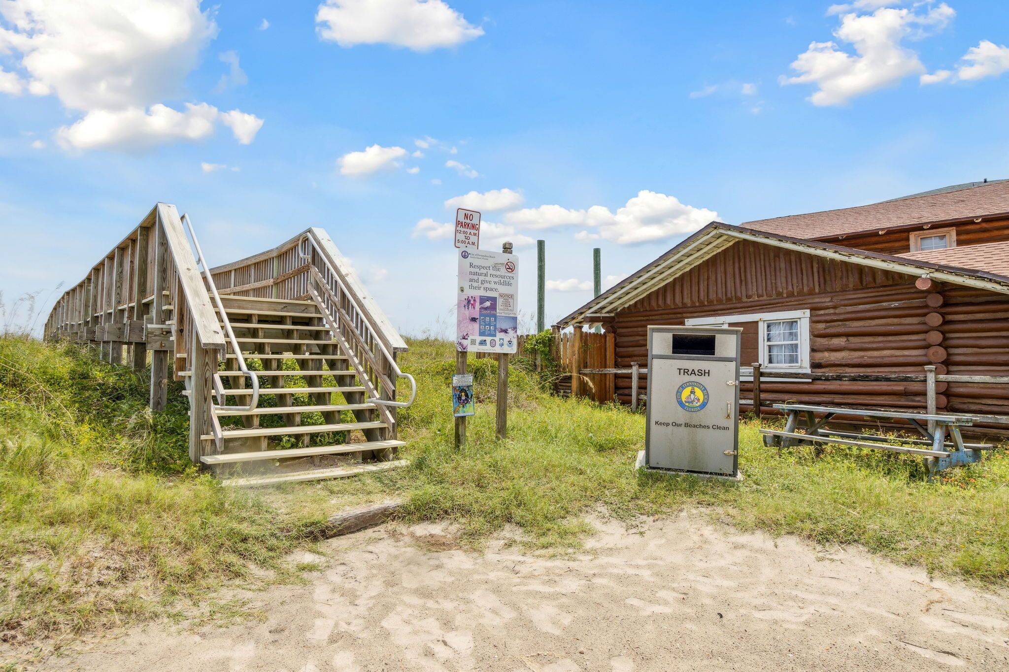 A rustic wooden building with a staircase leading up to a porch, a trash bin, and a signboard.