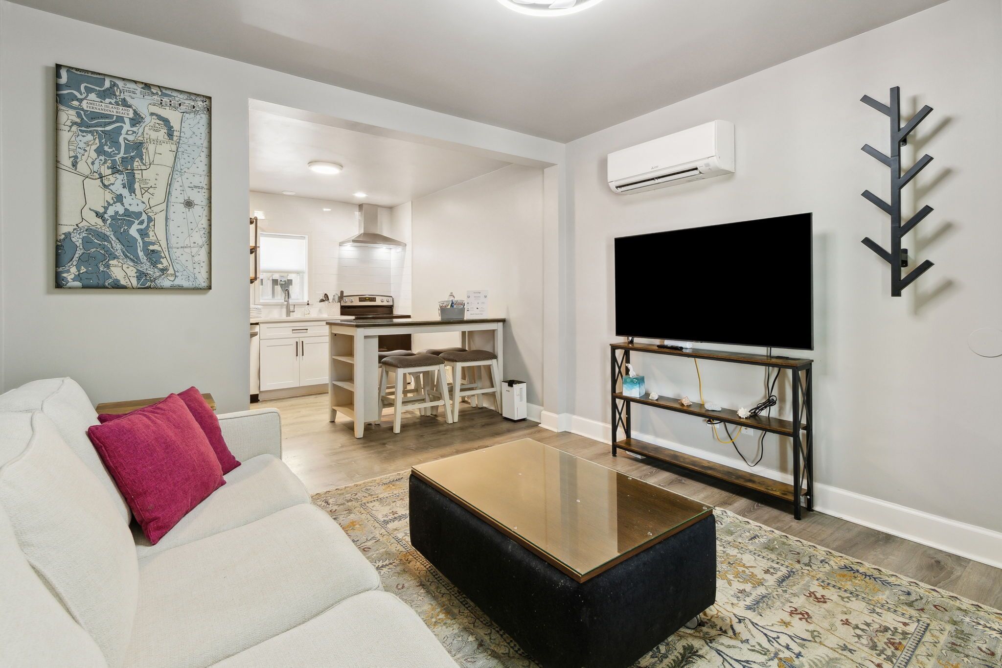 A modern living room with a sofa, coffee table, and a TV on a stand. There is a kitchen in the background with a dining area.
