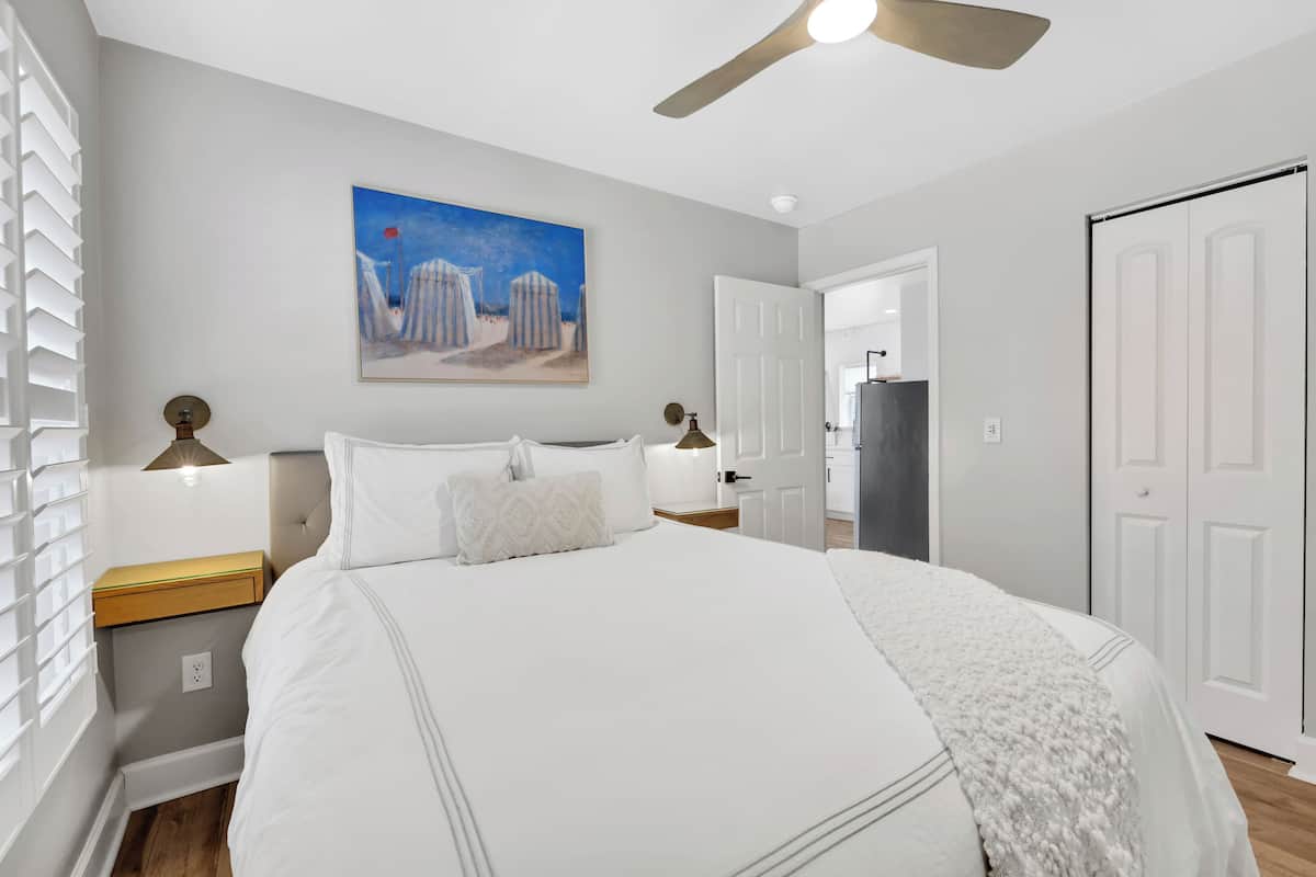 A bedroom with a large bed, a ceiling fan, a painting on the wall, and a door leading to another room.