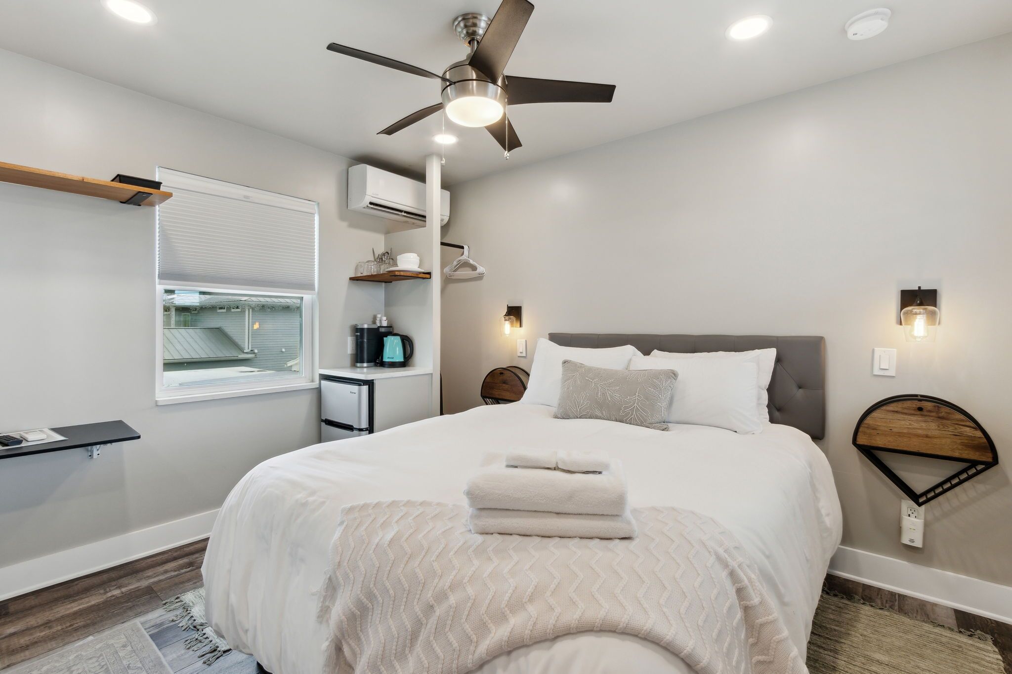 A modern bedroom with a large bed, a ceiling fan, a bedside table, a nightstand, a wall-mounted shelf, a window with blinds, and a wall-mounted light fixture.