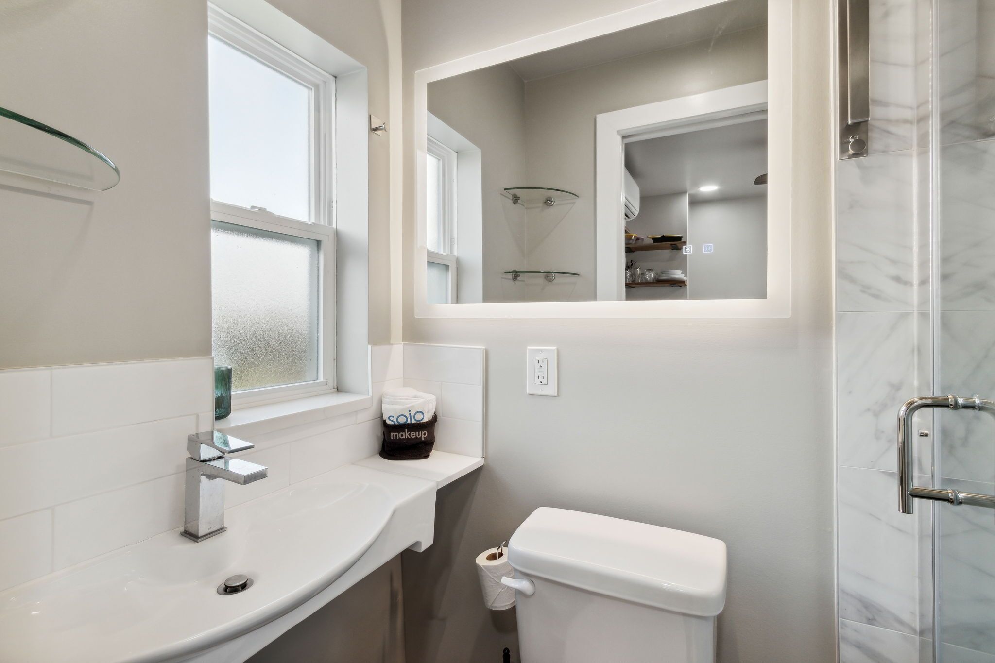A bathroom with a white sink, a glass shower enclosure, a toilet, and a mirror with a frosted window above.