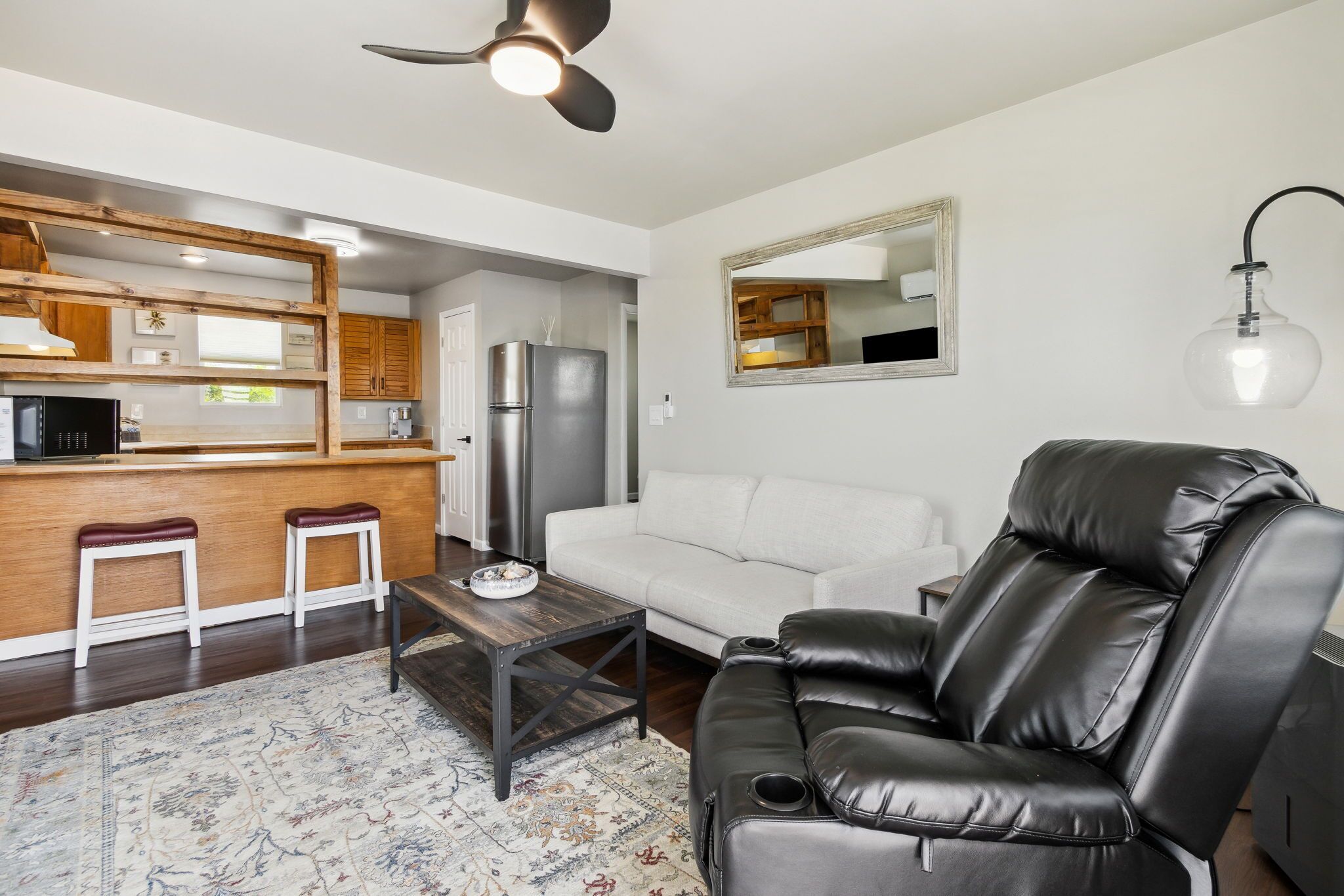 A modern living room with a sofa, a black leather recliner, a wooden coffee table, and a kitchen area with a refrigerator and wooden cabinets.