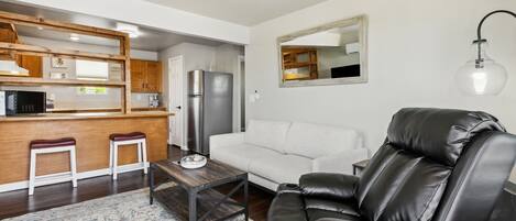 A modern living room with a sofa, a black leather recliner, a wooden coffee table, and a kitchen area with a refrigerator and wooden cabinets.