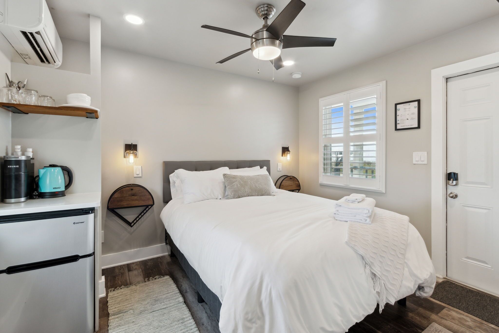 A bedroom with a bed, a ceiling fan, a window with blinds, a door, and a small kitchenette with a refrigerator and a kettle.
