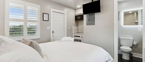 Un dormitorio con cama, ventilador de techo, televisor montado en la pared y baño con inodoro y lavamanos.