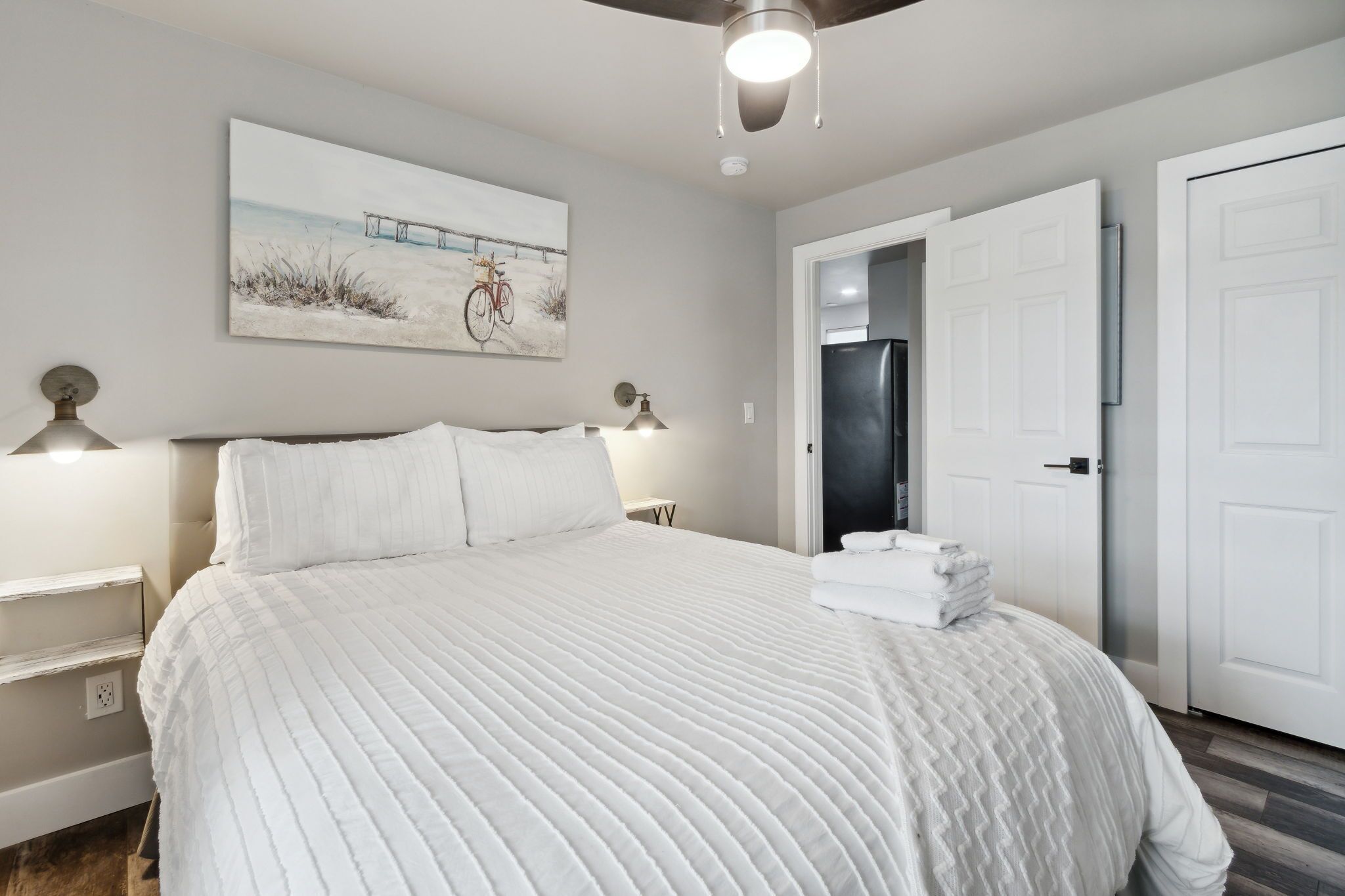 A neatly made bed with a white quilt, a bedside lamp, and a painting of a bicycle on a wall.
