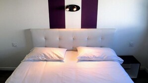 A neatly made bed with white linens and two pillows, a headboard with a light fixture, and a small bedside table. - Hôtel Le Rohu (Sene)