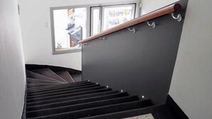 A staircase with a wooden handrail, a black carpeted landing, and a window with a view of the outside. - Hôtel Le Rohu (Sene)