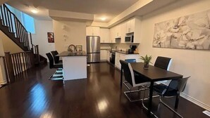 Una cocina moderna con electrodomésticos de acero inoxidable, un comedor con mesa y sillas negras, y una escalera que sube a otro nivel.