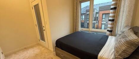 A bedroom with a single bed, a window with curtains, a door, and a carpeted floor.
