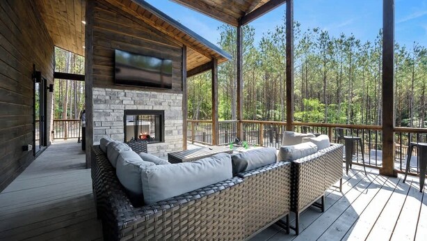 A covered deck with a stone fireplace, wicker furniture, and a view of a forest. - Pets | Heated Pool | Game Room | Sleeps 36 (Broken Bow)