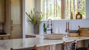 A kitchen with wooden cabinets, a marble countertop, a sink, a potted plant, a cactus, and a window with a view of trees.